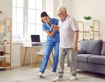 Young nurse helping elderly man walk in the room, holding his hand, supporting him. Treatment and rehabilitation after injury or stroke, life in assisted living facility, senior care concept.